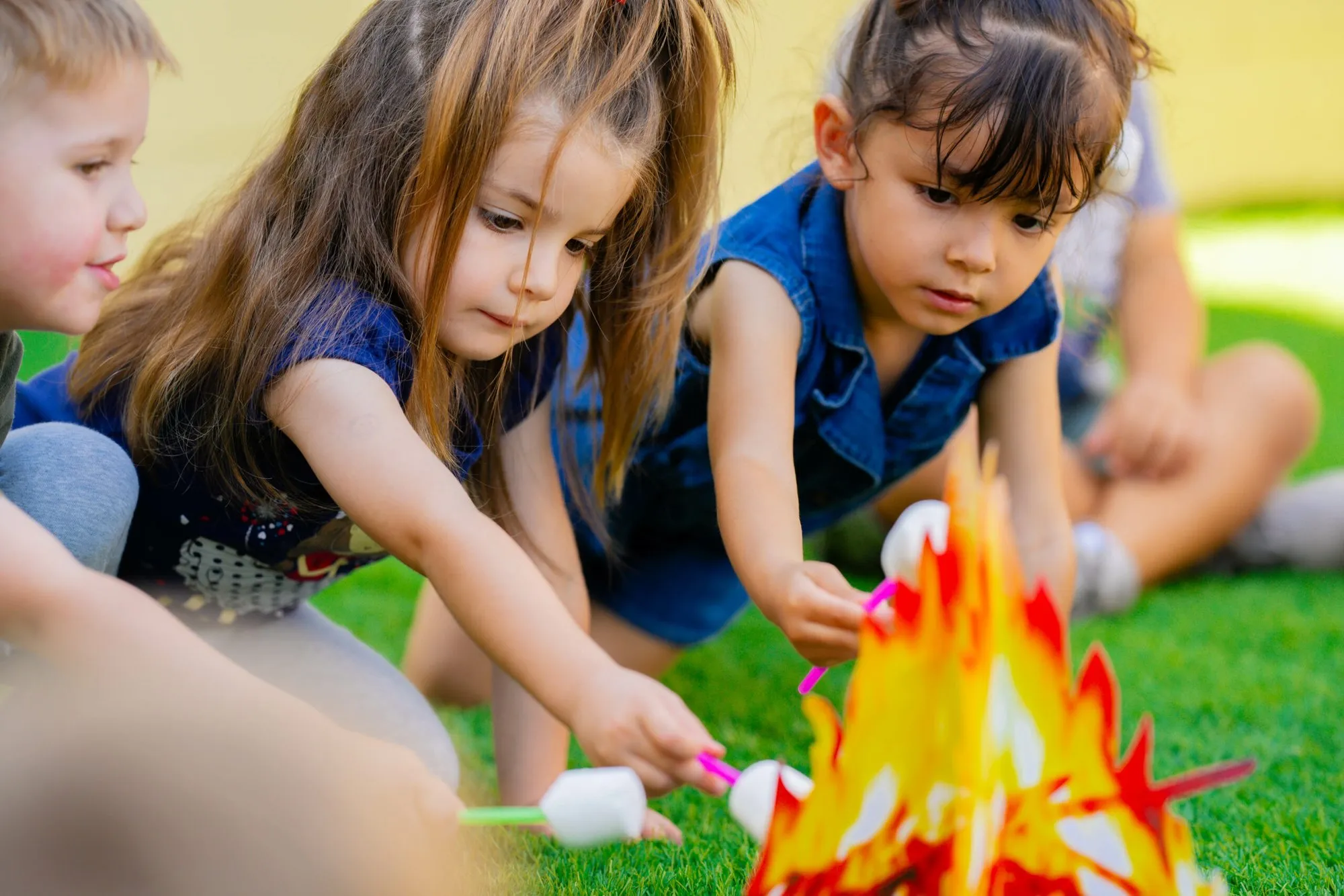 Two girls kneel on the grass and hold pretend marshmallows over a paper campfire during outdoor play.