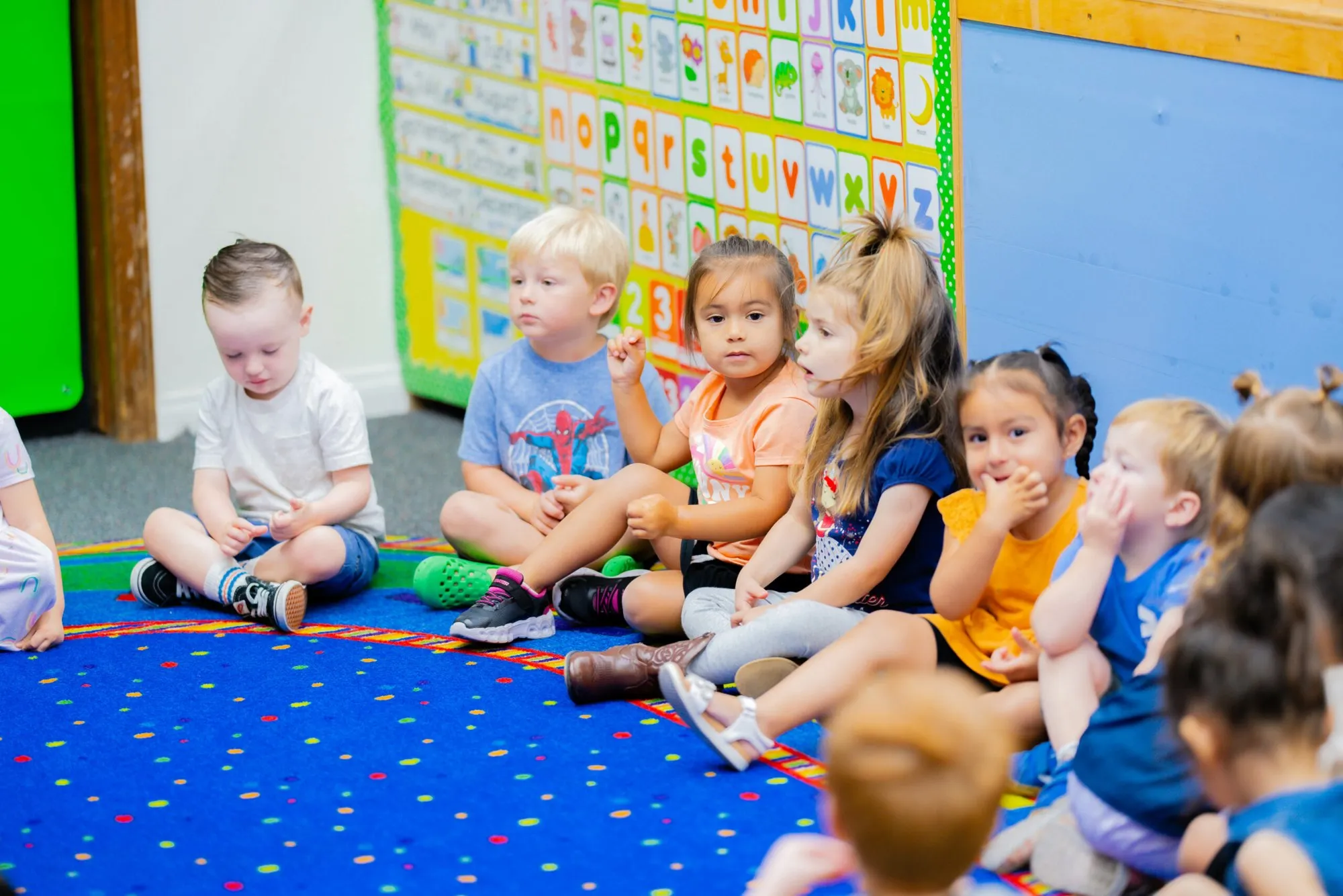 Preschool children sit together on a blue rug during circle time in a bright classroom.