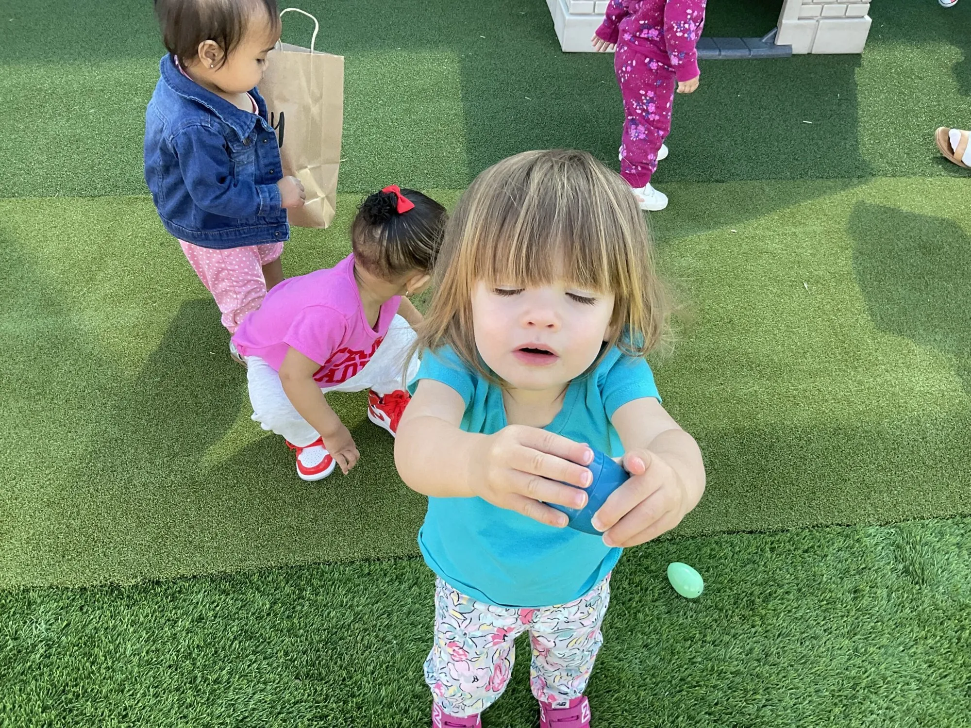A toddler on the lawn holds out a blue egg she found during an outdoor egg hunt.