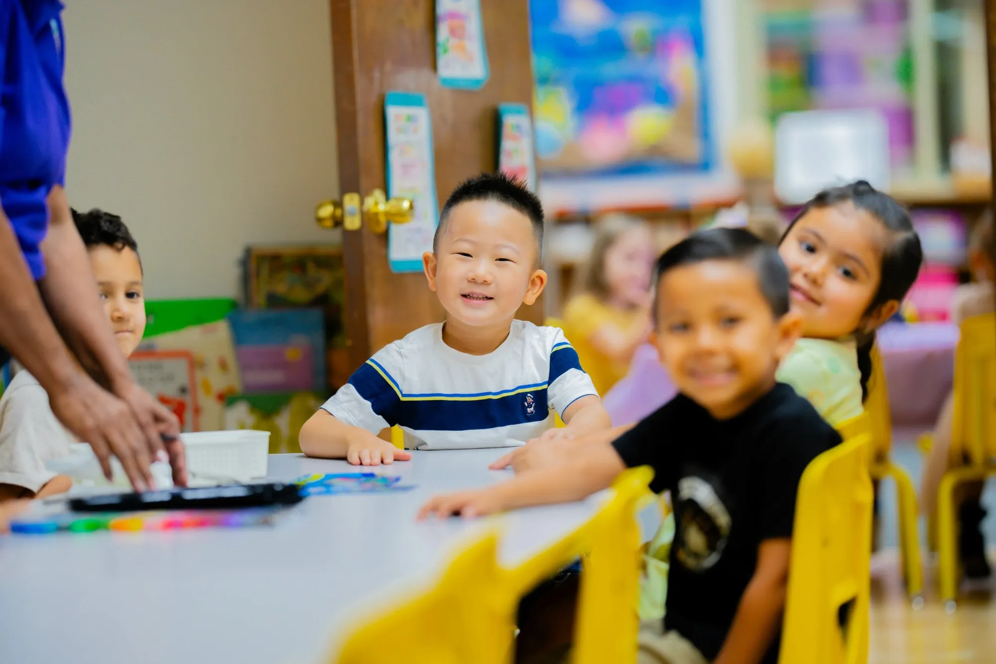 Smiling children look up from their classroom table while a teacher sets out materials.
