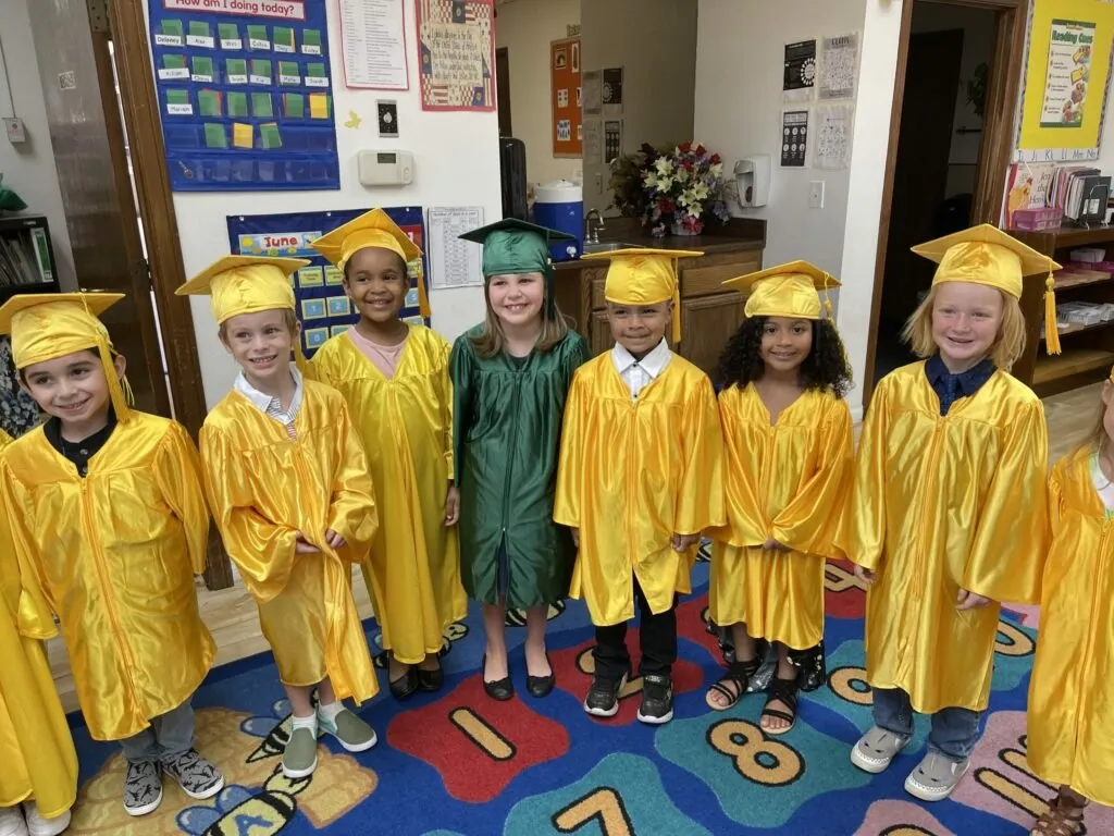 Kindergarten graduates in gold and green caps and gowns stand together in their classroom.