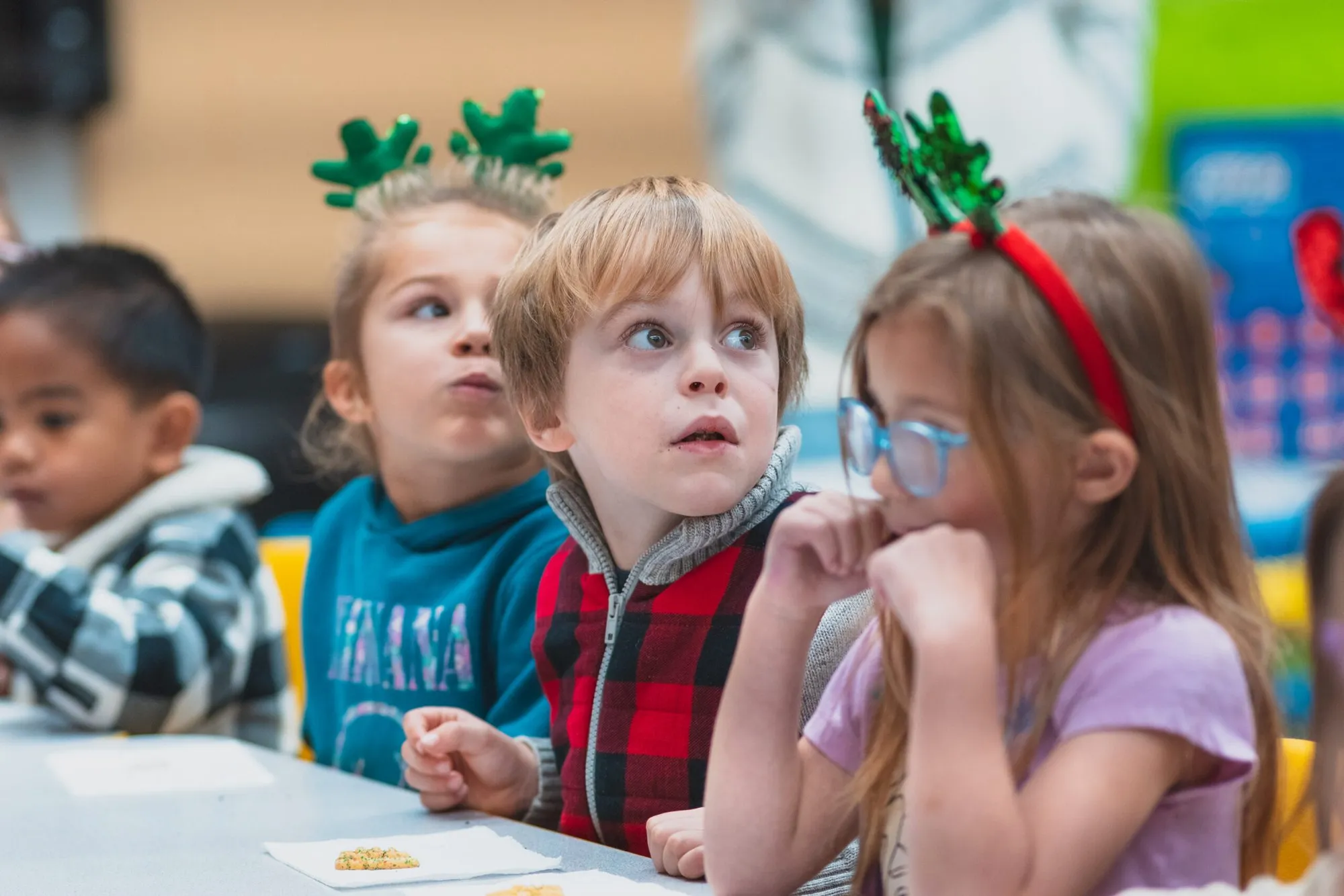 Four children in festive headbands sit at a table, looking up with curiosity.