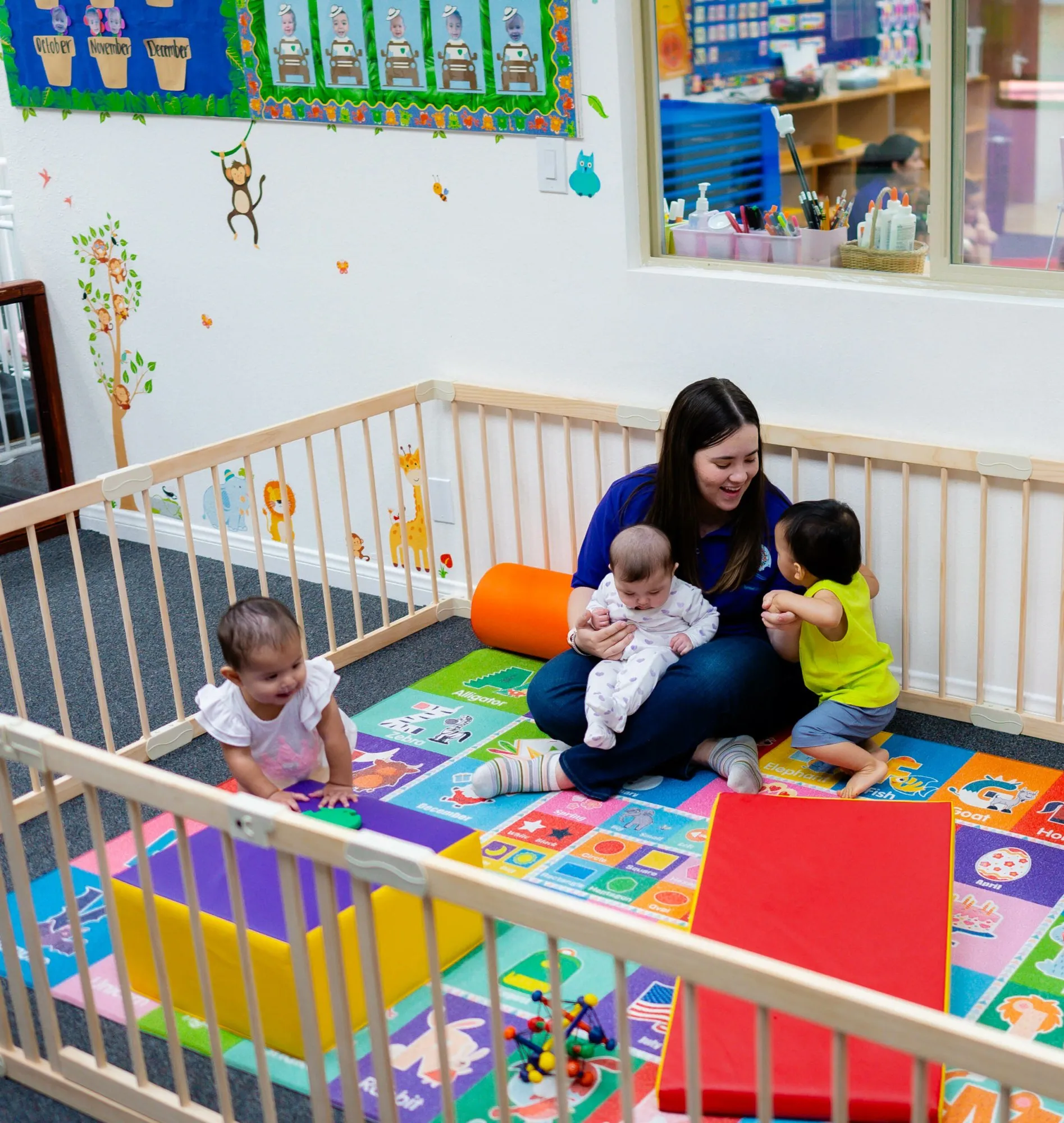 A teacher sits on a colorful play mat holding a baby while two toddlers play beside her in the infant room.