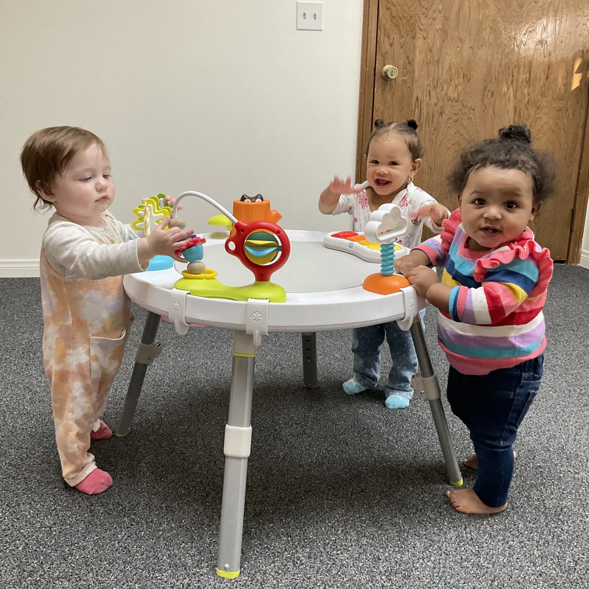 Three babies stand around a low activity table exploring toys together.