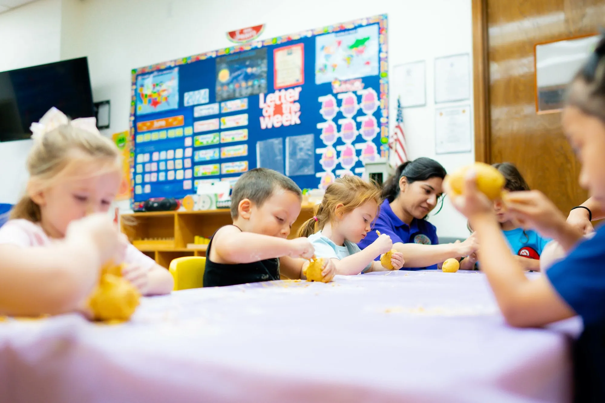 Children squeeze lemons at a classroom table while a teacher helps a classmate beside them.