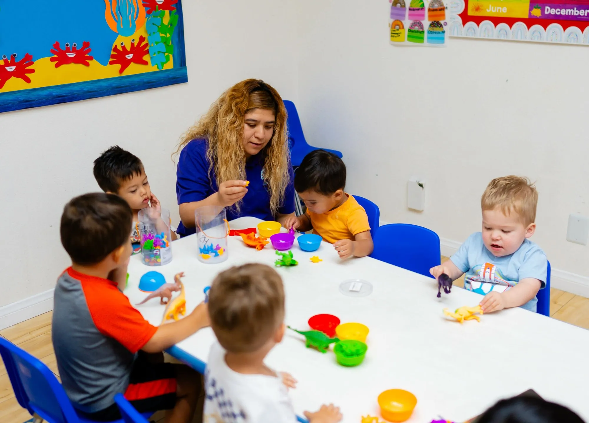 A teacher sits at a table with toddlers working with colorful sorting cups.